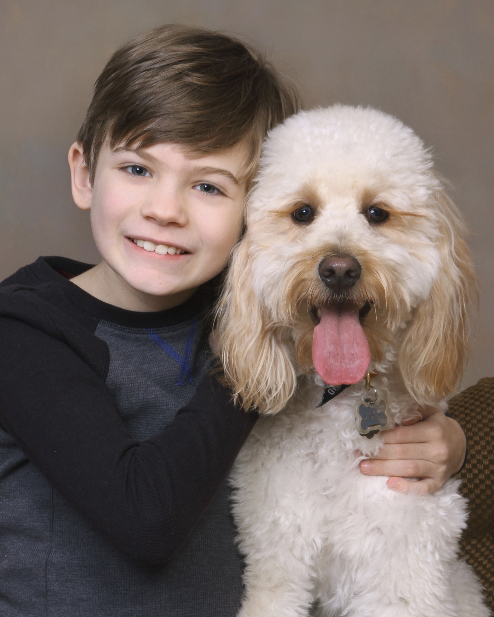 Young boy with brunette hair, smiling with his arms wrapped around his poodle mix