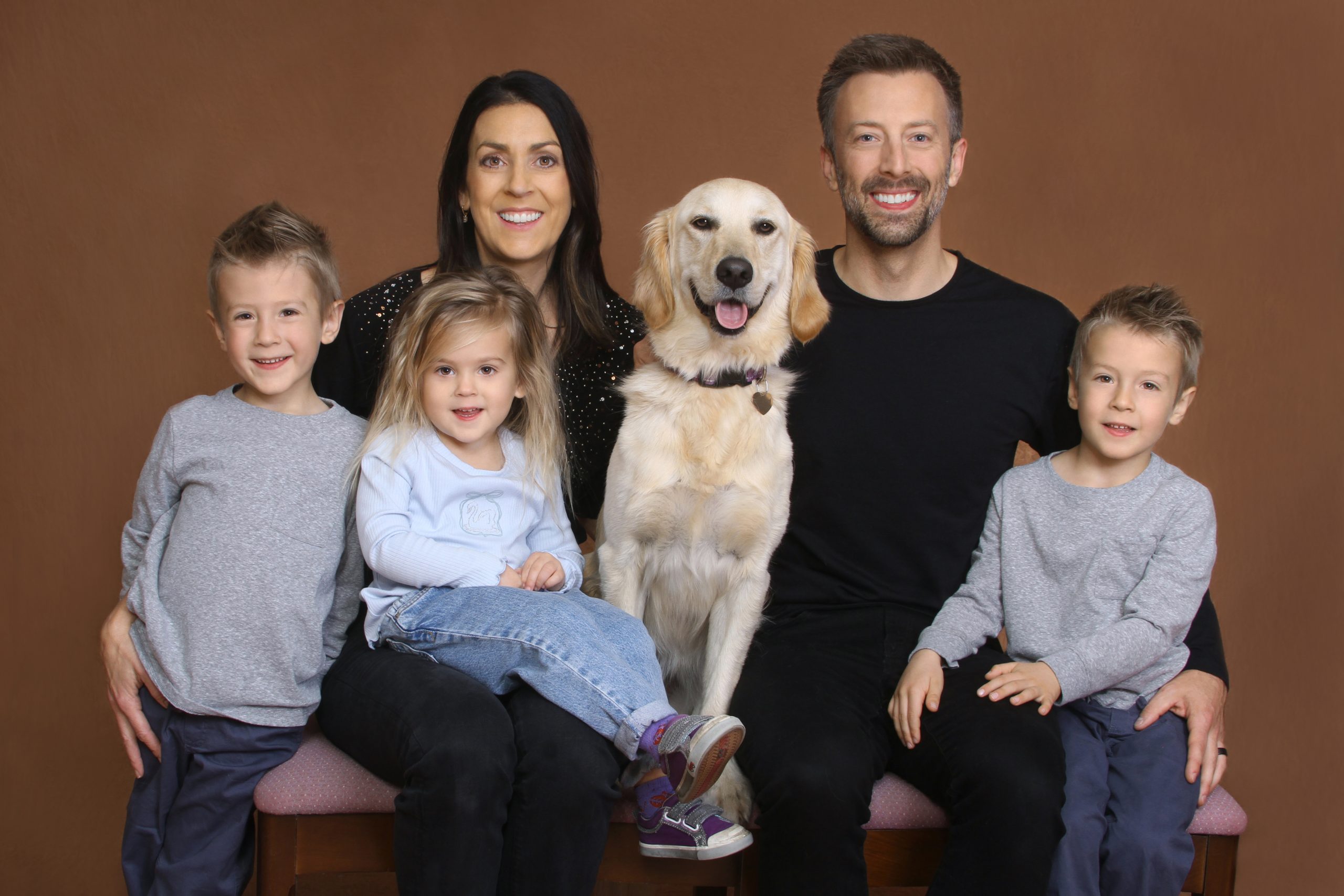 Tan background of a portrait studio, family of four including a chestnut golden retriever, smile for the camera.